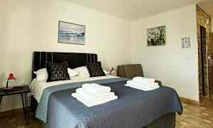 Ett hotellrum med en säng täckt med ett blått överkast, två kuddar och hopvikta handdukar ovanpå. Tavlor hänger på väggarna, och ett sängbord med en röd lampa står bredvid sängen. (AI-genererad text)