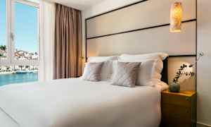 Hotellrum med en dubbelsäng, prydnadskuddar och ett nattduksbord. Utsikt över vatten och byggnader genom fönstret. (AI-genererad text)