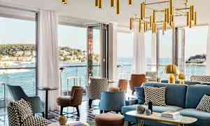 En modern lounge med sittmöbler och dekorativa ljuskronor, omgiven av stora fönster som erbjuder utsikt över en kuststad och havet. (AI-genererad text)