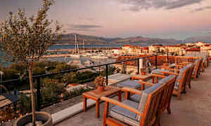 Uteservering med trästolar och bord, utsikt mot småbåtshamn och berg i solnedgång. (AI-genererad text)