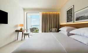 Hotellrum med bäddad säng, skrivbord, stol och utsikt mot havet. (AI-genererad text)