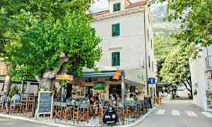 Ett utomhuscafé vid en byggnad med vita väggar och gröna fönsterluckor, omgivet av grönska och träd. (AI-genererad text)