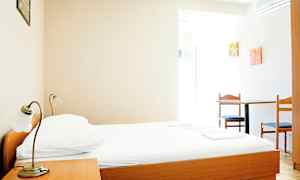 Ett enkelt hotellrum med en säng av trä, ett sängbord, och ett bord med två stolar. (AI-genererad text)