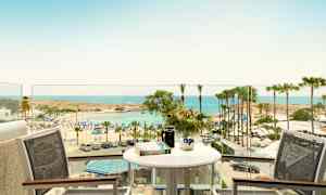 Vy från en balkong med bord och stolar mot en strand med parasoller och palmer vid havet. (AI-genererad text)