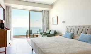 Hotellrum med säng, sittgrupp och utsikt mot havet genom glasdörrar. (AI-genererad text)