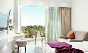 Ett ljust hotellrum med en soffa, en säng med lila täcke och en balkong som har utsikt över grönska. (AI-genererad text)