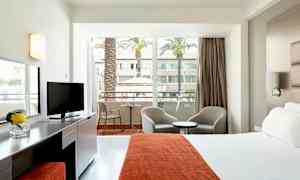 Hotellrum med säng, sittplatser och balkong som vetter mot en innergård med palmer. (AI-genererad text)