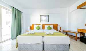 Två enkelsängar med grön överkast och vita kuddar i ett hotellrum med ljus inredning. (AI-genererad text)