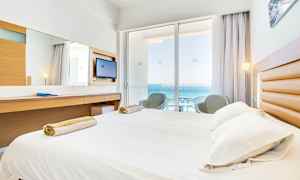 Hotellrum med säng, skrivbord och balkong med utsikt över havet. (AI-genererad text)