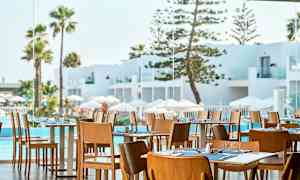 Restaurang med trästolar och dukade bord, utsikt mot vit arkitektur, pool och palmer. (AI-genererad text)