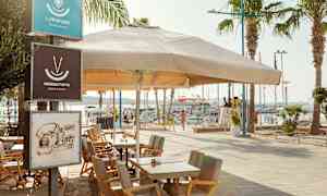 Utomhusservering med bord och stolar under ett stort parasoll vid en hamn. (AI-genererad text)