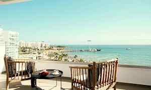 Två trästolar och ett bord på ett hotellbalkong med utsikt över strand och hav. (AI-genererad text)