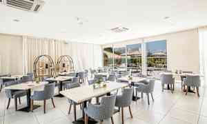 Ett ljust och modernt restaurangområde med runda och fyrkantiga bord omgivna av stoppade stolar. Stora fönster visar en uteservering. (AI-genererad text)