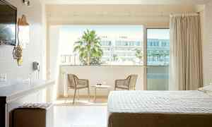 Ett hotellrum med en säng, skrivbord och stol bredvid en balkong med utsikt över stadsmiljö. (AI-genererad text)