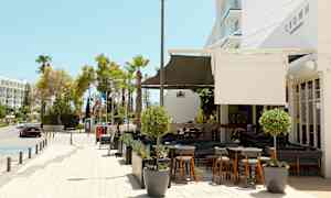 Bild av en utomhusrestaurang och trottoar intill en gata med träd och byggnader i bakgrunden. (AI-genererad text)