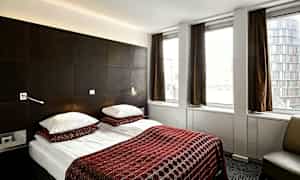 Ett hotellrum med en dubbelsäng bäddad med rödmönstrade överkast och två kuddar. Rummet har mörka möbler och tre stora fönster med bruna gardiner. (AI-genererad text)