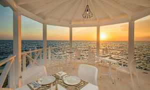 En utomhusrestaurang med vita möbler, belägen vid havet, med solnedgång i bakgrunden. (AI-genererad text)