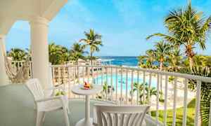 På bilden syns en avkopplande balkong med bord och stolar, samt en hammock. Utsikten är över en tropisk pool omgiven av palmer och havet i bakgrunden. (AI-genererad text)