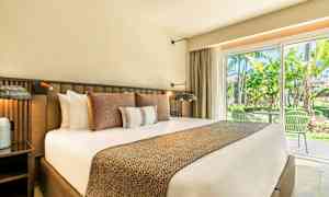 Ett hotellrum med en stor säng utrustad med flera kuddar, sängbord och en glasad dörr som leder till en uteplats och gröna omgivningar. (AI-genererad text)