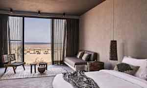 Ett hotellrum med säng, soffa och utsikt mot stranden genom en stor glasdörr. (AI-genererad text)