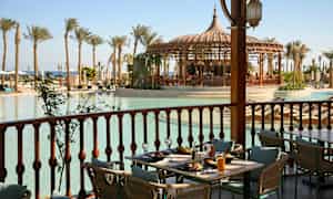 Ett uppdukat bord på en altan med utsikt över en pool omgiven av palmer och en paviljong. (AI-genererad text)