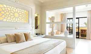 Ett ljust och elegant sovrum med dekorerade vaser på en hylla, en stor säng med kuddar och utgång till en balkong. (AI-genererad text)