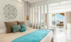 Ett hotellrum med ljusa väggar, en dekorerad säng med blå och beige sängkläder, samt en balkong med havsutsikt. (AI-genererad text)