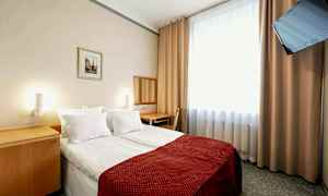 Ett hotellrum med en dubbelsäng, rödfärgad sängöverkast. En spegel och skrivbord står intill väggen. Ett fönster med ljusa gardiner och en väggmonterad TV finns i rummet. (AI-genererad text)