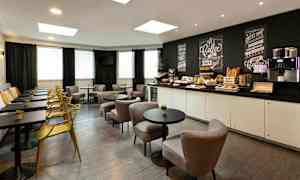 Ett caféområde med bord, stolar och en bänk längs väggen. Ett serveringsbord med frukosttillbehör och en kaffemaskin syns i bakgrunden. (AI-genererad text)