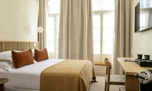 Hotellrum med en dubbelsäng, kuddar och en sängfilt i bruna toner samt ett skrivbord med en kaffemaskin. (AI-genererad text)