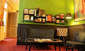 Ett rum med gröna väggar och eklektisk inredning, inklusive en hylla med böcker och dekorationer, en soffa, och en schackmönstrad stol. (AI-genererad text)