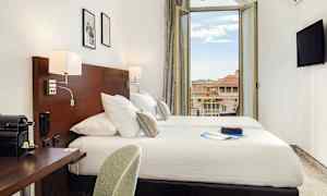 Ett hotellrum med dubbelsäng, skrivbord och en öppnad balkongdörr som visar utsikt över omgivande byggnader. (AI-genererad text)