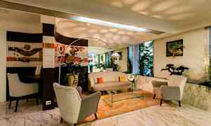 Ett vardagsrum med mjuka fåtöljer, bord och soffa på ett orange matt, med växter och konst på väggarna. Stor spegel och fönster. (AI-genererad text)