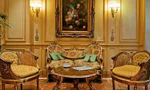 Ett elegant inrett rum med en pråligt dekorerad soffa, två matchande fåtöljer och ett soffbord framför. Oljemålning på väggen ovanför soffan. (AI-genererad text)