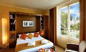 Ett hotellrum med en dubbelsäng, kuddar och filtar i varma färger samt en stor fönstervy mot träd och byggnader. (AI-genererad text)