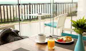 Frukostbord med kaffe, juice och grönsaker nära ett glasfönster med utsikt över en terrass med havet i bakgrunden. (AI-genererad text)