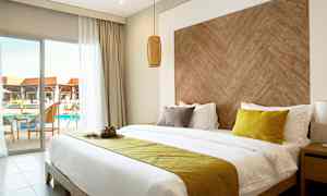 Hotellrum med dubbelsäng, gula och bruna kuddar, träinspirerad huvudgavel, samt balkong med utsikt över poolen. (AI-genererad text)