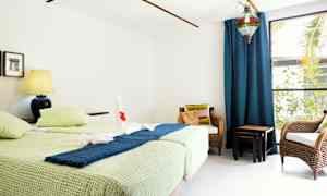 Ett rum med två enkelsängar, en blå lampa på ett sängbord, två rottingstolar och ett stort fönster med blå gardiner. (AI-genererad text)