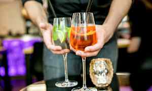 Två drinkar hålls fram av en person, ett glas innehåller en orange cocktail och det andra en klar cocktail med lime och mynta. (AI-genererad text)