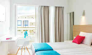 Ett ljust hotellrum med två enkelsängar, vita och turkosa sängkläder, en rosa kudde. Fönstret har ljusa gardiner och öppnar mot balkonger. (AI-genererad text)