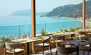 En restaurang med uteservering och utsikt över ett turkosblått hav och kuperad kustlinje. (AI-genererad text)