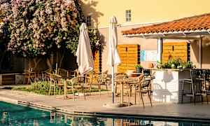 Utomhusterrass med bord och stolar vid en pool, omgiven av växter och ett träd med blommor. (AI-genererad text)