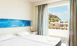 Ett hotellrum med två enkelsängar, ljusa väggar och en blå dekorativ väggpanel. Det finns en stor fönsteröppning med utsikt över en kulle och byggnader utanför. (AI-genererad text)