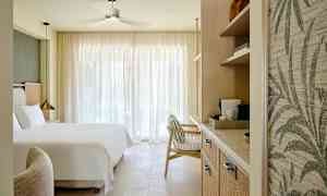 Ett hotellrum med en stor säng, en fåtölj och hyllor med kaffemaskin, belyst av naturligt ljus genom stora gardiner. (AI-genererad text)