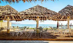 Utomhusområde med bord och stolar under halmtak nära stranden. (AI-genererad text)