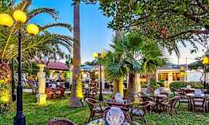 En utomhusrestaurang med rottingbord och stolar, palmer och dekorerade ljuslyktor, omgiven av grönska och belysning. (AI-genererad text)