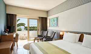 Ett hotellrum med en dubbelsäng, två stolar, ett bord och en tv-möbel. En balkong med utsikt mot palmer är synlig genom en glasdörr. (AI-genererad text)