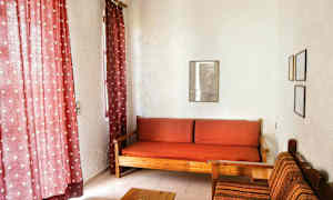 Vardagsrummet har röda gardiner, en träsitssoffa med rödt karerade kuddar, och en tygklädd stol bredvid. (AI-genererad text)