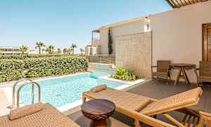 Bild föreställer en terrass med solstolar, ett litet bord och två stolar bredvid en privat pool. I bakgrunden syns grönskande buskar och palmer. (AI-genererad text)