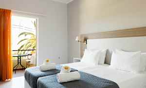 Ett hotellrum med två enkelsängar med grå överkast och vita kuddar samt handdukar dekorerade med gula blommor. Ett fönster med orange gardiner öppnar mot en balkong med utsikt över palmer. (AI-genererad text)